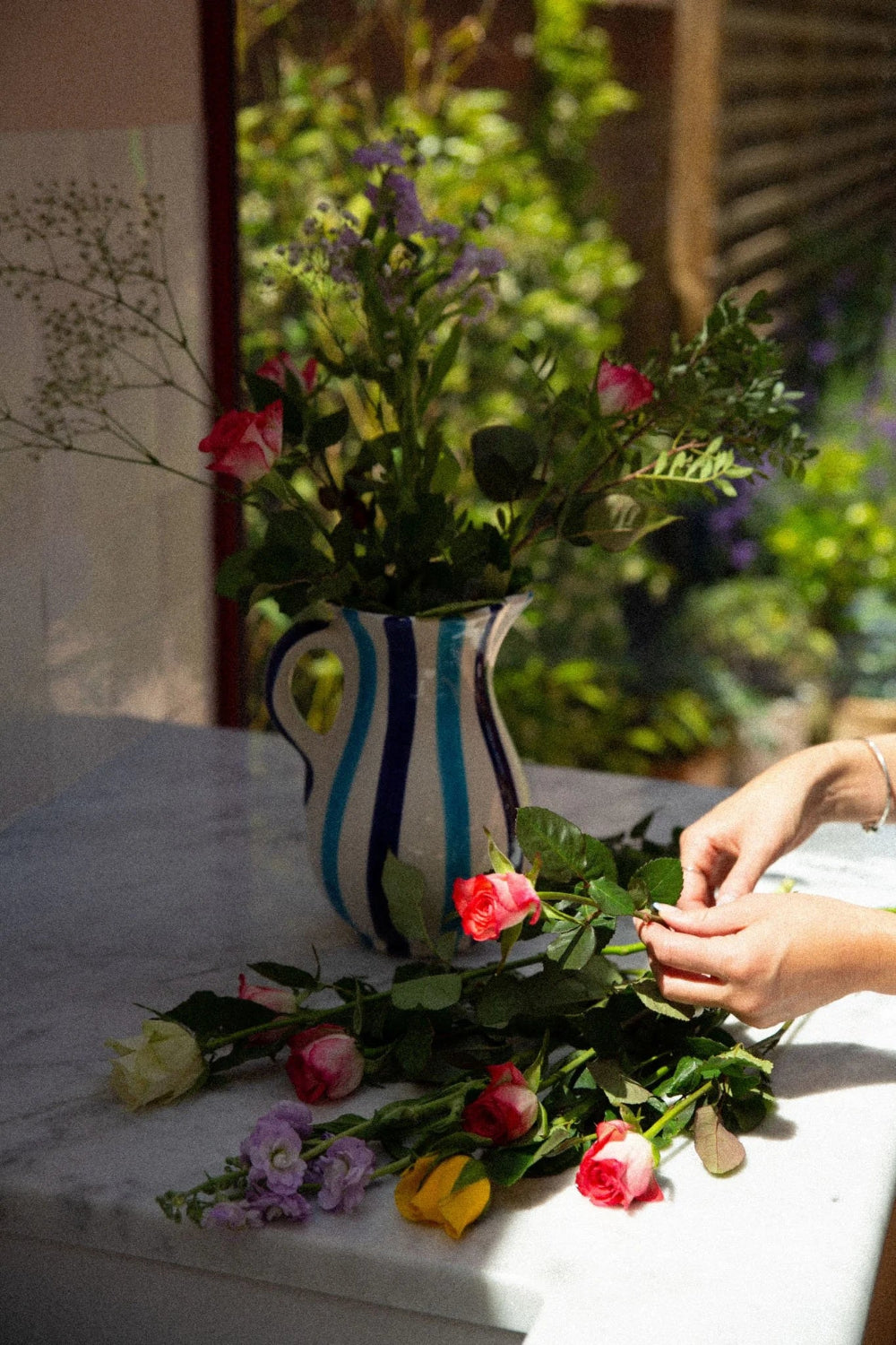 Person arranging flowers on a table with a blue striped ceramic mug by woven rosa in the background