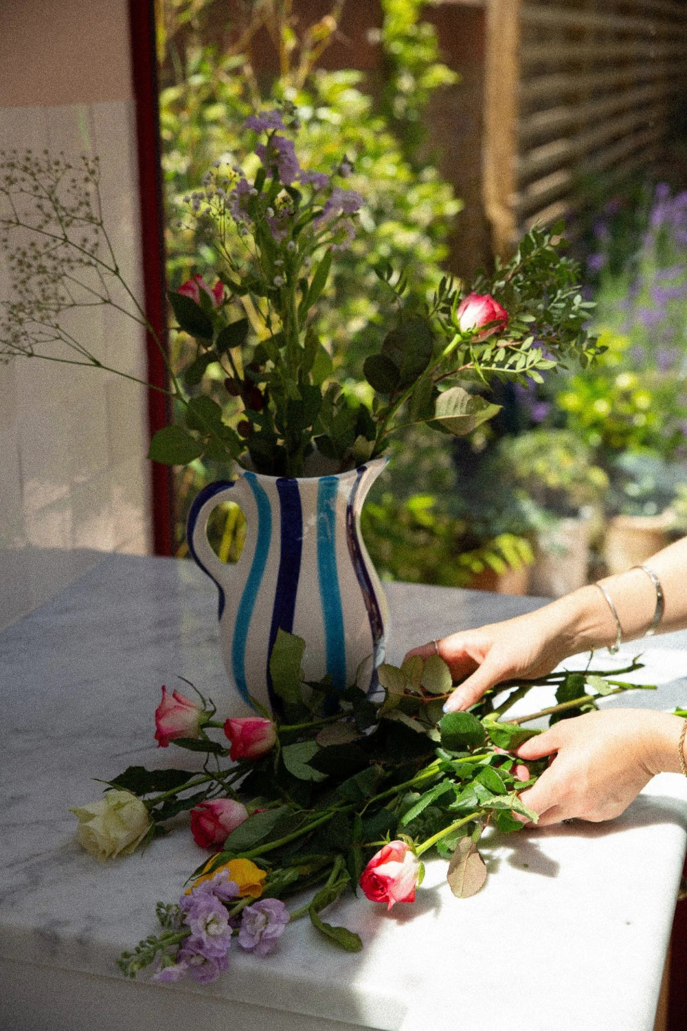 A striped ceramic pitcher filled with mixed flowers, placed on a white marble table outdoors, with a person's hands arranging additional roses and flowers nearby.