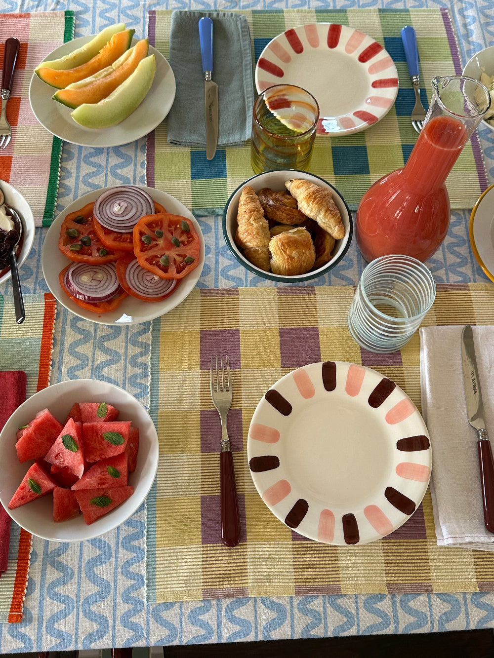 A colorful breakfast table setting with patterned plates, a glass bottle of orange juice, bowls of sliced watermelon, tomatoes, red onion, and croissants, and a striped table runner.