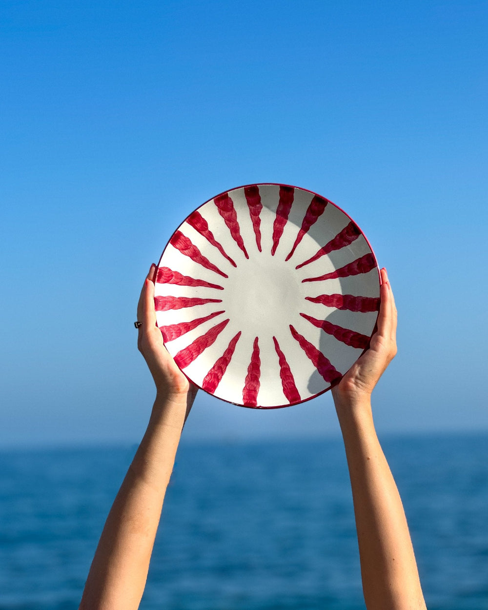 A person holds a round white plate with a red sunburst pattern above the ocean, with a clear blue sky in the background.