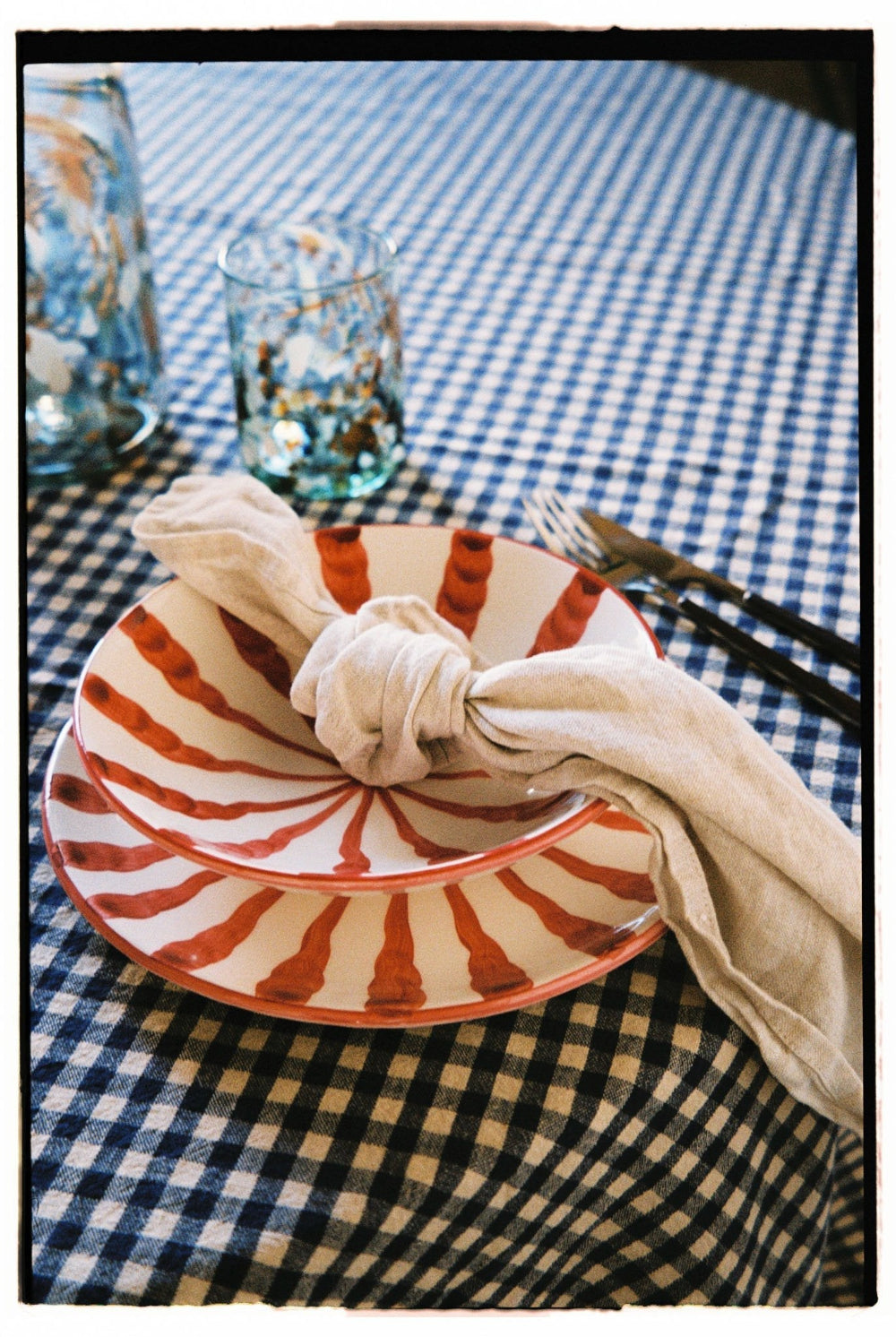 A place setting on a blue and white gingham tablecloth, featuring a stack of white plates with a red-orange sunburst pattern, a beige cloth napkin tied in a knot, and silver cutlery.
