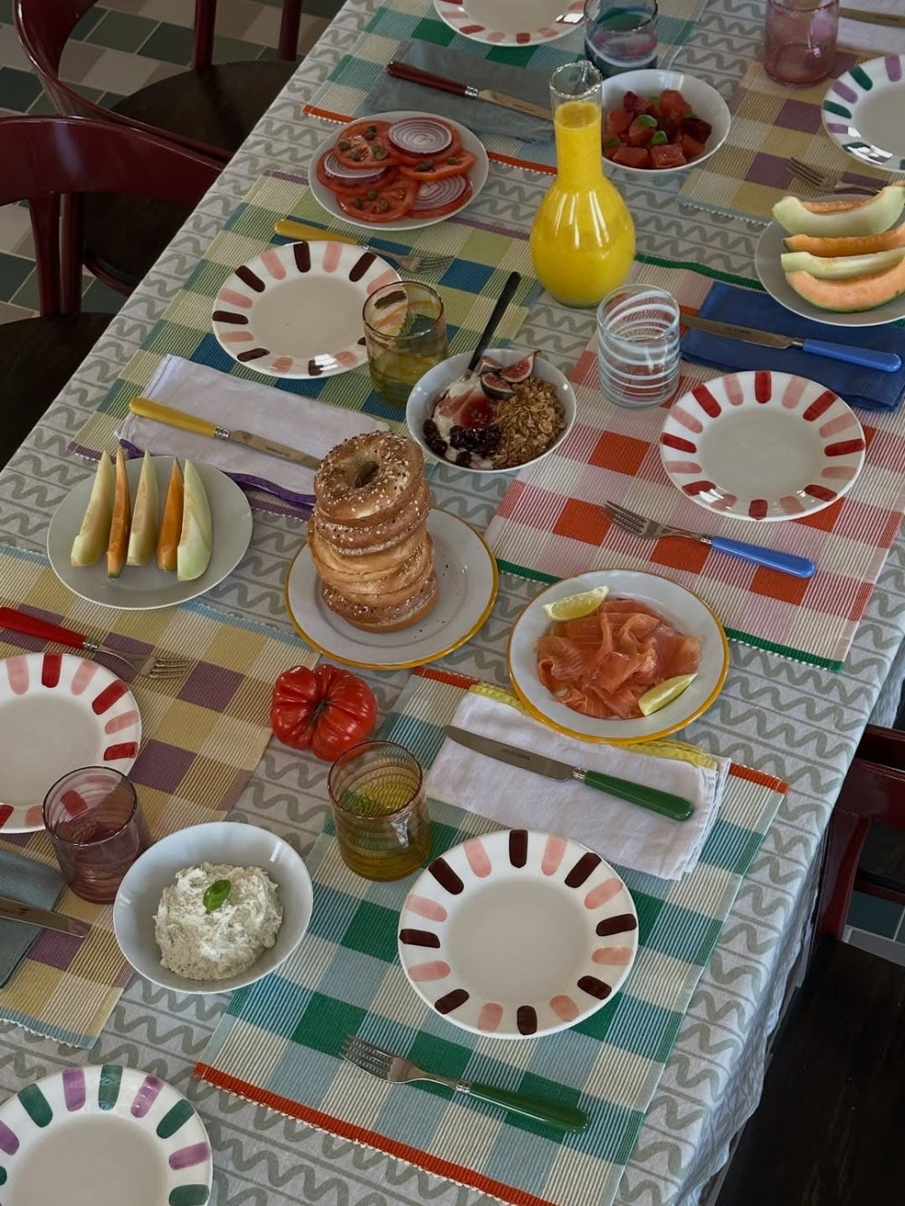A styled breakfast table setting with colorful patterned plates, bowls of food including bagels, tomatoes, strawberries, and a yellow juice bottle, all arranged on a checkered tablecloth.