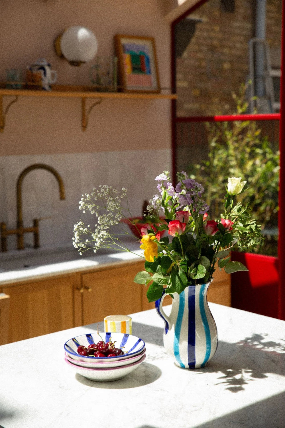 A striped ceramic pitcher filled with a colorful bouquet of wildflowers sits on a white marble countertop in a sunlit kitchen. A matching striped bowl with cherries is also visible nearby.