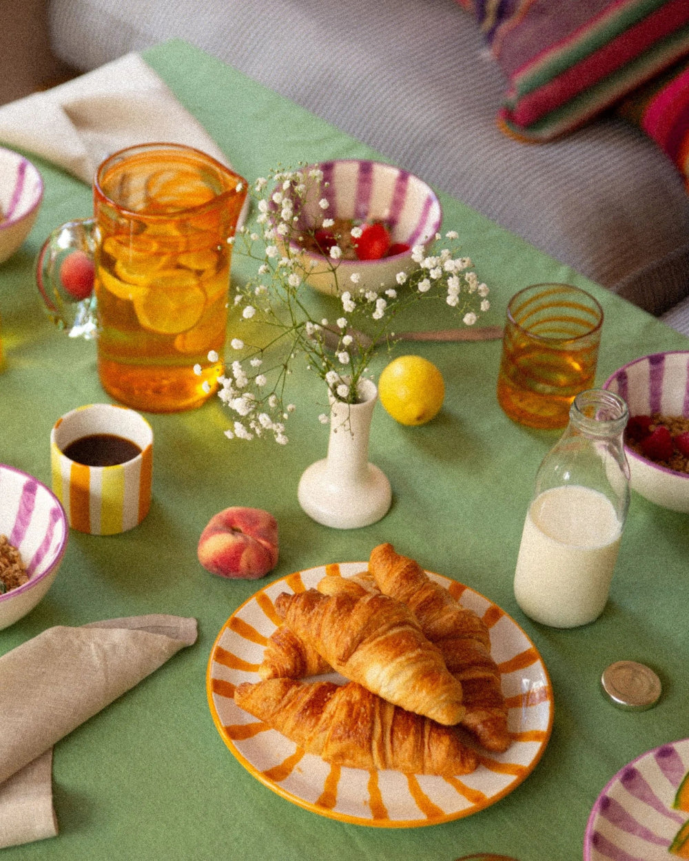 A styled breakfast table scene with croissants on a striped plate, a glass bottle of milk, a pitcher of iced tea with lemon slices, small bowls, a vase of baby's breath, and fruit on a green tablecloth.