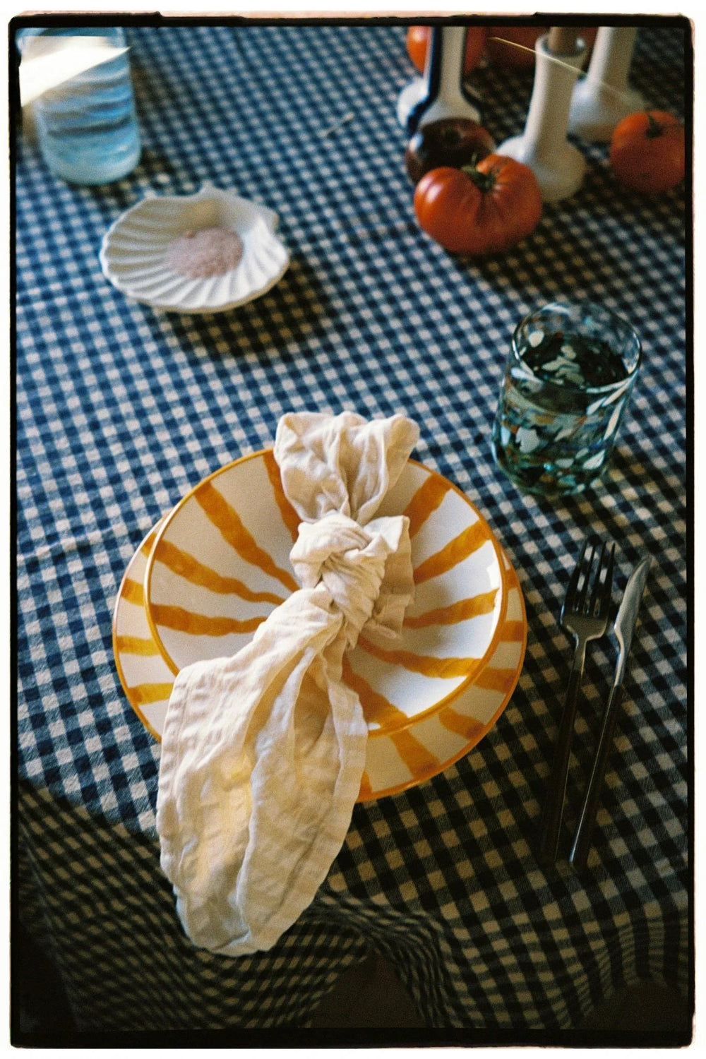 A styled tabletop setting with a blue-and-white checkered tablecloth, a striped yellow and white plate, a cream fabric napkin tied in a knot, black cutlery, and small autumn decor items in the background.