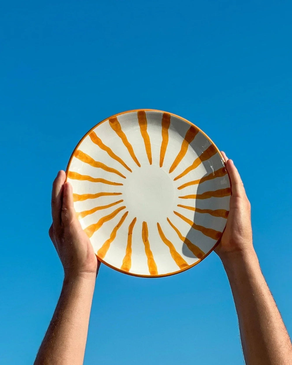 A ceramic plate with a white center and orange radial stripe pattern, held up by two hands against a clear blue sky.
