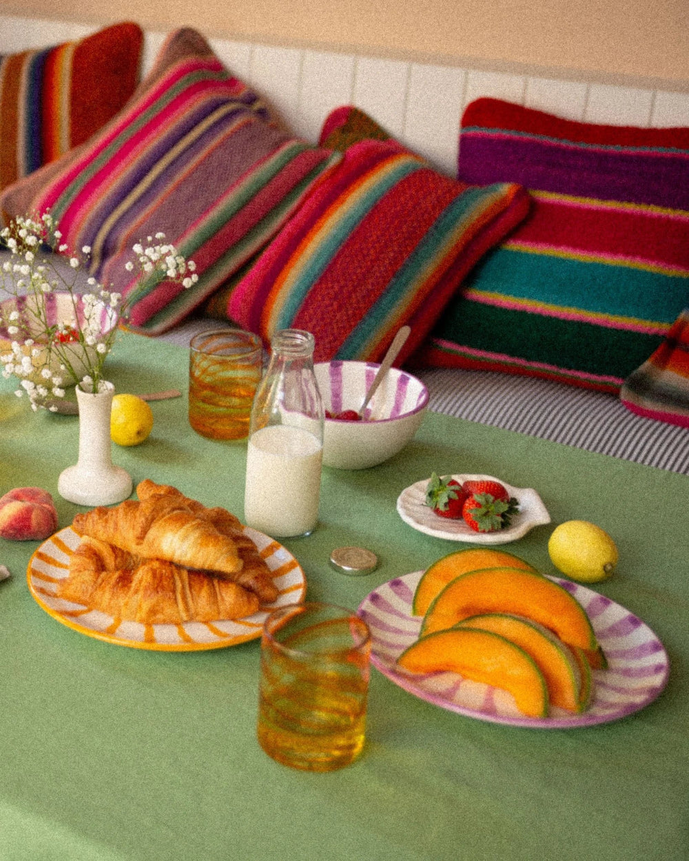 A styled breakfast table scene with croissants on a striped plate, sliced melon on a pink-and-white striped plate, a glass bottle of milk, a bowl of fruit, lemons, and small flowers, set in front of colorful striped pillows on a sofa.