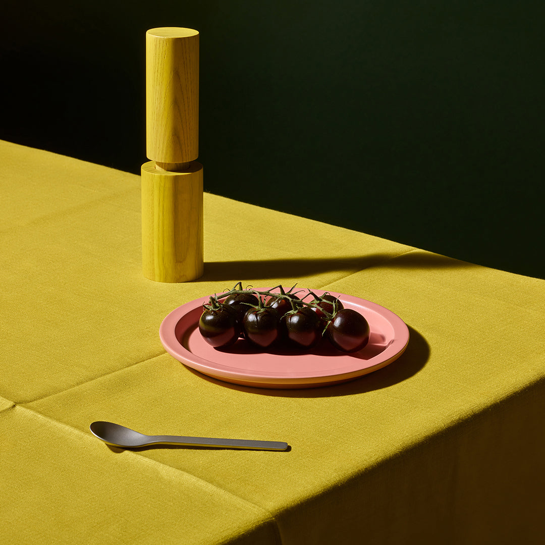 A pink ceramic plate holding a cluster of dark cherry tomatoes on a yellow tablecloth, with a wooden cylindrical salt or pepper mill and a metal spoon nearby in a styled tabletop scene.