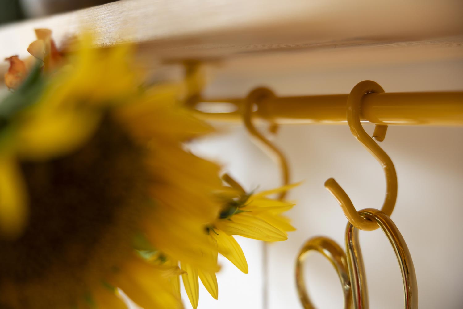Close-up of a yellow kitchen rail rack rod with decorative S-shaped hooks, partially obscured by blurred yellow flowers in the foreground.