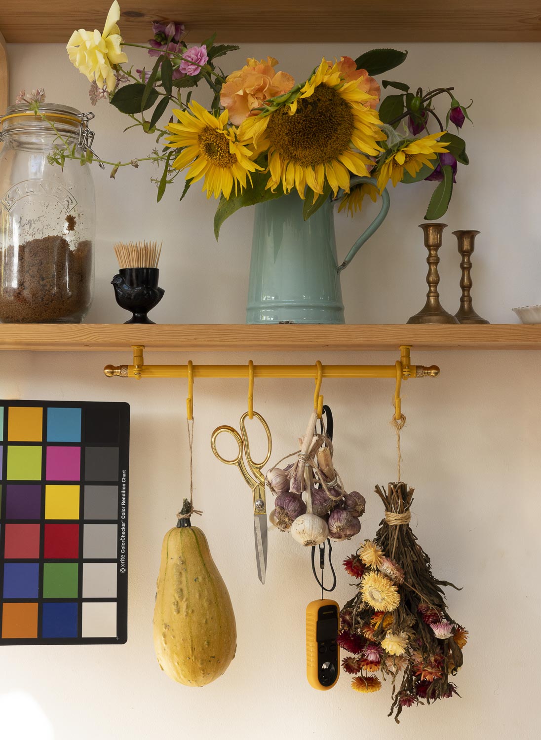 A rustic wall-mounted wooden shelf display with a mint green pitcher of sunflowers, a glass jar, brass candlesticks, and a yellow hanging rod holding scissors, garlic, dried flowers, and a squash on a cream wall.
